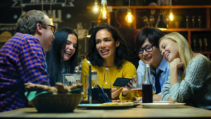 Frau zeigt ihren Freunden interessante Sachen auf ihrem Smartphone, während sie in der Bar gute Zeit haben. Sie lachen, scherzen, trinken in Hipster-Bar.