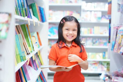 Asian children or kid girl red shirt happy smile and shopping or choose tale or story book and read on bookshelf in bookstore or library room at kindergarten school or nursery for learn and study