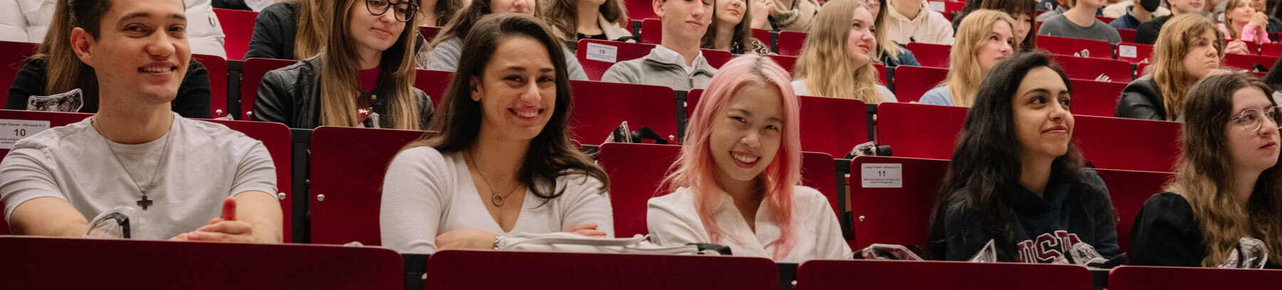 Erstsemester-Studentinnen und Studenten der FAU sitzen im Hörsaal. Einige im Vordergrund lächeln in die Kamera. Der Rest schaut in Richtung der Professorin oder des Professors. 