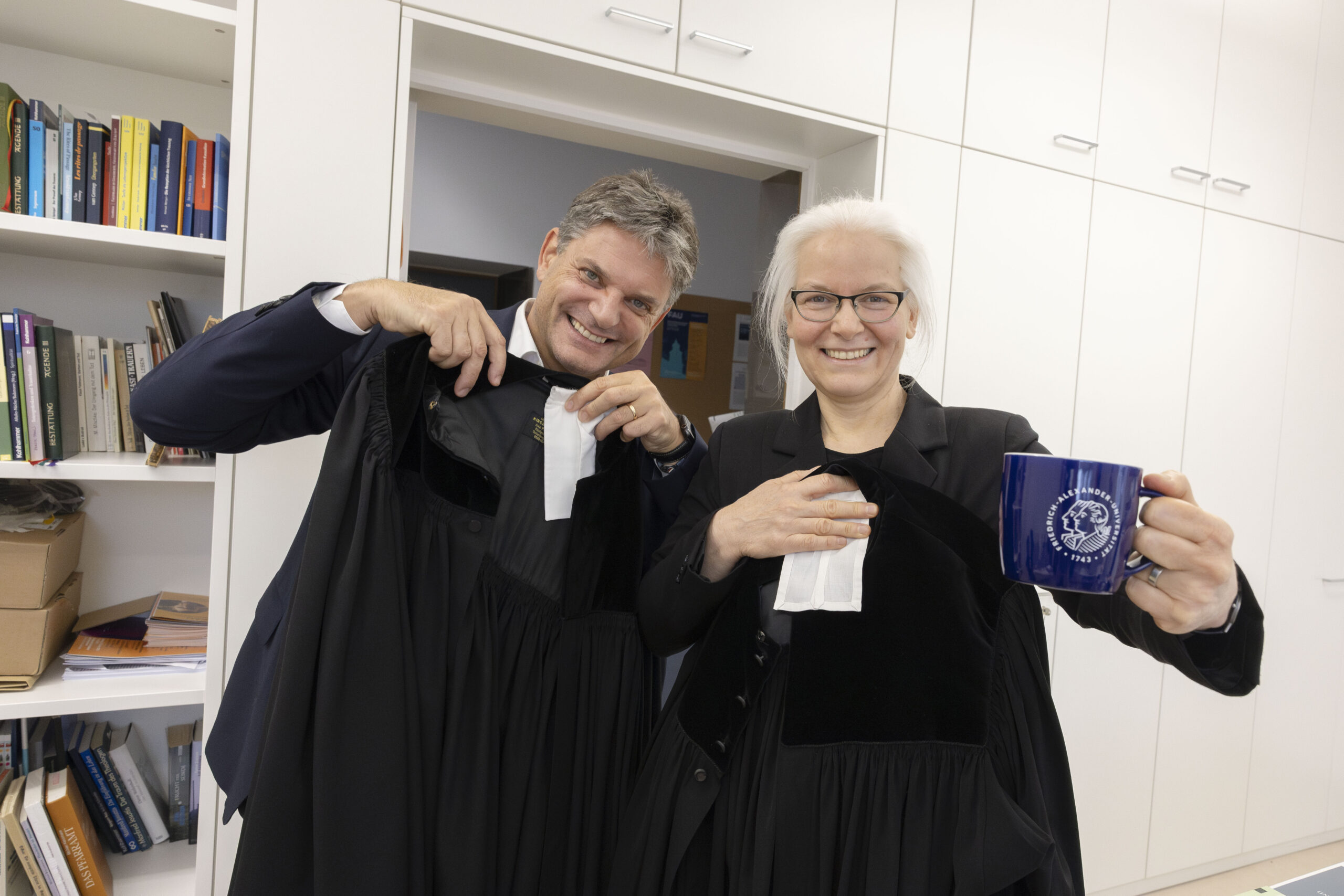 Eine Frau und ein Mann lächeln in die Kamera. Die Frau hält eine FAU-Tasse in der Hand. 