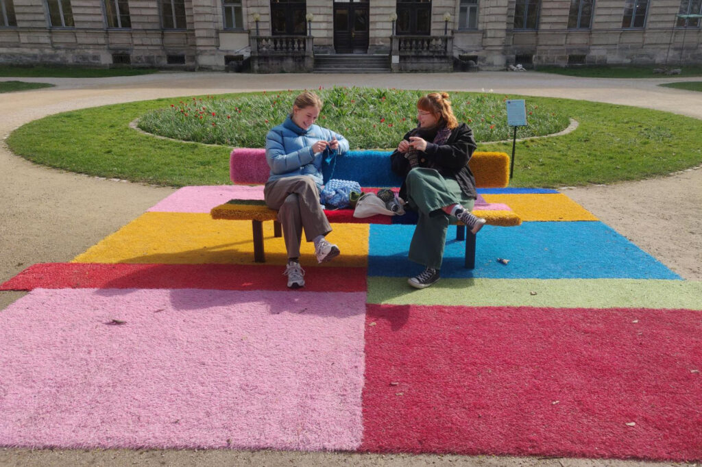 Kunstwerk der Ausstellung „Beyond Boundaries“ zeigt eine bunte Parkbank mit Teppich. Auf ihr sitzen zwei Studentinnen und Stricken.