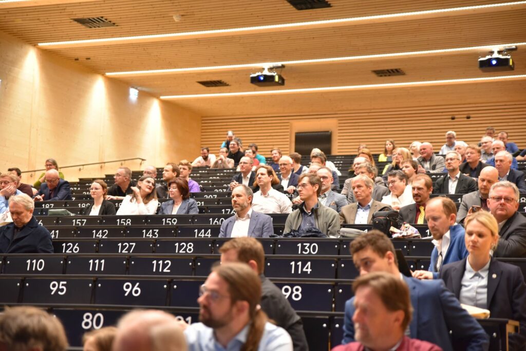 Hörsaal von unten am Podium in den Raum hinein fotografiert. Man sieht ansteigende Stuhlreihen zum Teil belegt von Menschen.