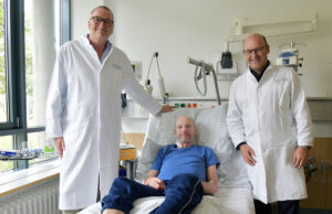 Zwei Arzte stehen neben einem Krankenhausbett auf dem ein Patient liegt. Sie lächeln in die Kamera.