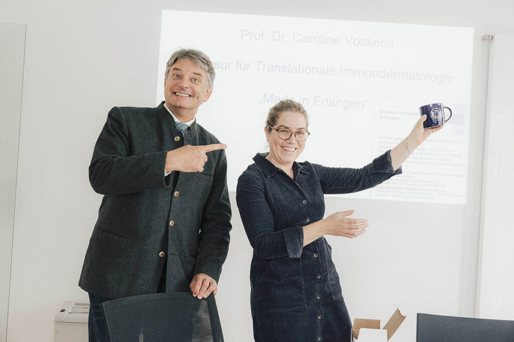 Voskens holds up an FAU mug and Hornegger points at Voskens, who is standing next to him. 