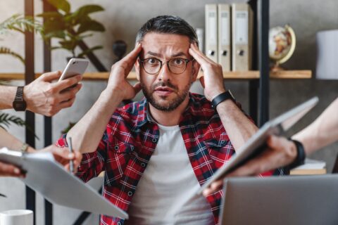 Überarbeiteter Mann schaut in Kamera während ihm verschiedene Tablets und Handys ins Gesicht gehalten werden.