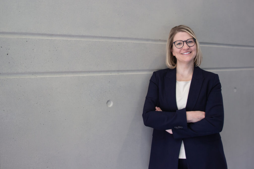 Dr. Gensberger Reigl, eine Frau mit Brille, und schulterlangen Haaren, im schwarzern Sacko, lehnt mit verschränkten Armen an einer Wand und lächelt.