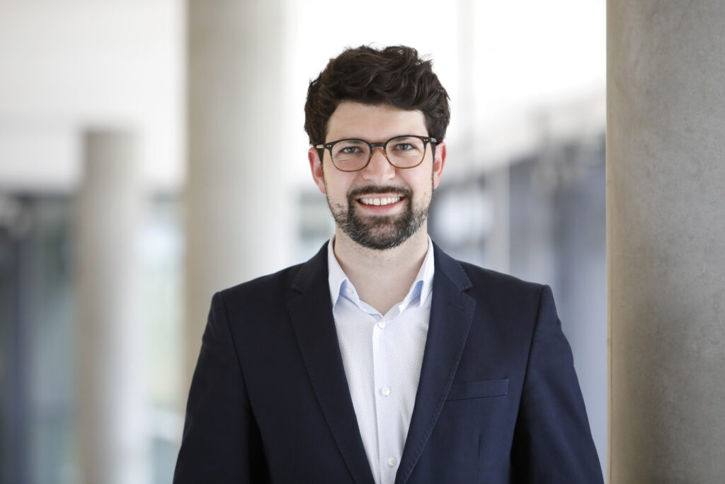Adrian Meier, ein junger mann mit Brille, dunklem Bart und dunklem, vollem Haar.