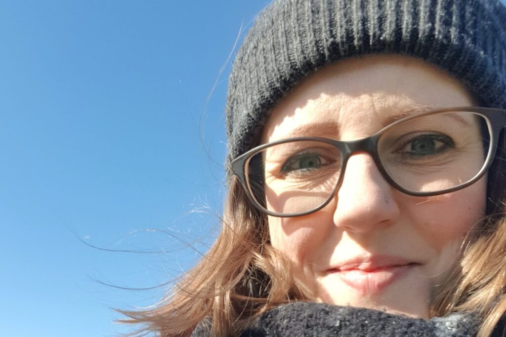Portrait von FAU-Alumna Heike Nowak-Schwerdtfeger vor einem SChrifstzug der Digital Tech Academy. Sie trägt eine dunkelrahmige Brille und hat die langen braunen Haare zu einem Zopf gebunden. Sie lächelt freundlich in die Kamera.