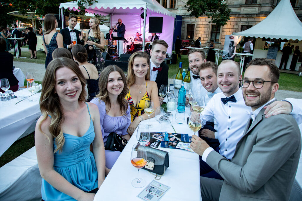 Gruppe junger Studierender die auf dem Schlossgartenfest zusammensitzen und in die Kamera lächeln. Im Hintergrund ist ein Band zu sehen.
