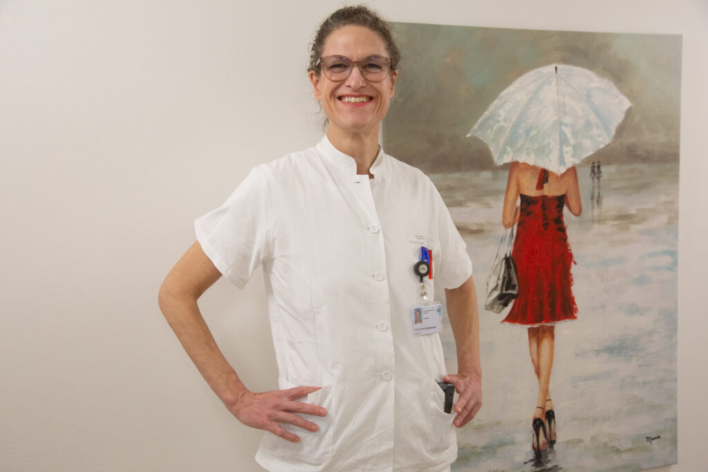Anne Schützen ist ein Frau mittleren Alters mit lockigem Haar und einer Brille. Sie trägt einen weißen arztkittel, kim hintergrund hängt ein bild mit Frau im roten Kleid.