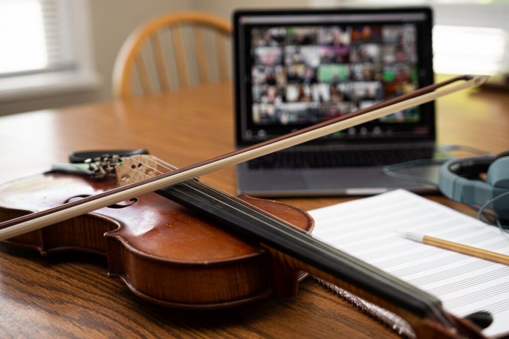 Eine Geige und ein Laptop auf einem Tisch. Auf dem Laptop sind verschwommen Bildkacheln von musizierenden Menschen in einem Online-Meeting zu sehen.