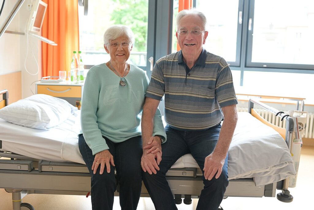 Das Ehepaar P. sitzt gemeinsam auf einen Krankenhausbett.