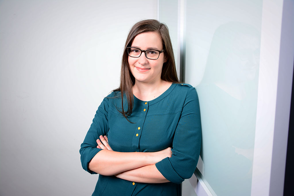 Ein Bewerbungsfoto von Dr. Julia Hauser: Der Hintergrund ist weiß und sie lehnt sich an eine Glaßwand.