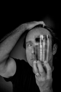 Schwarz-weiß-Foto von Bjarne Mädel: Es ist nur die linke Gesichtshälfte zu sehen, da er sich vor die rechte ein Wasserglaß hält.