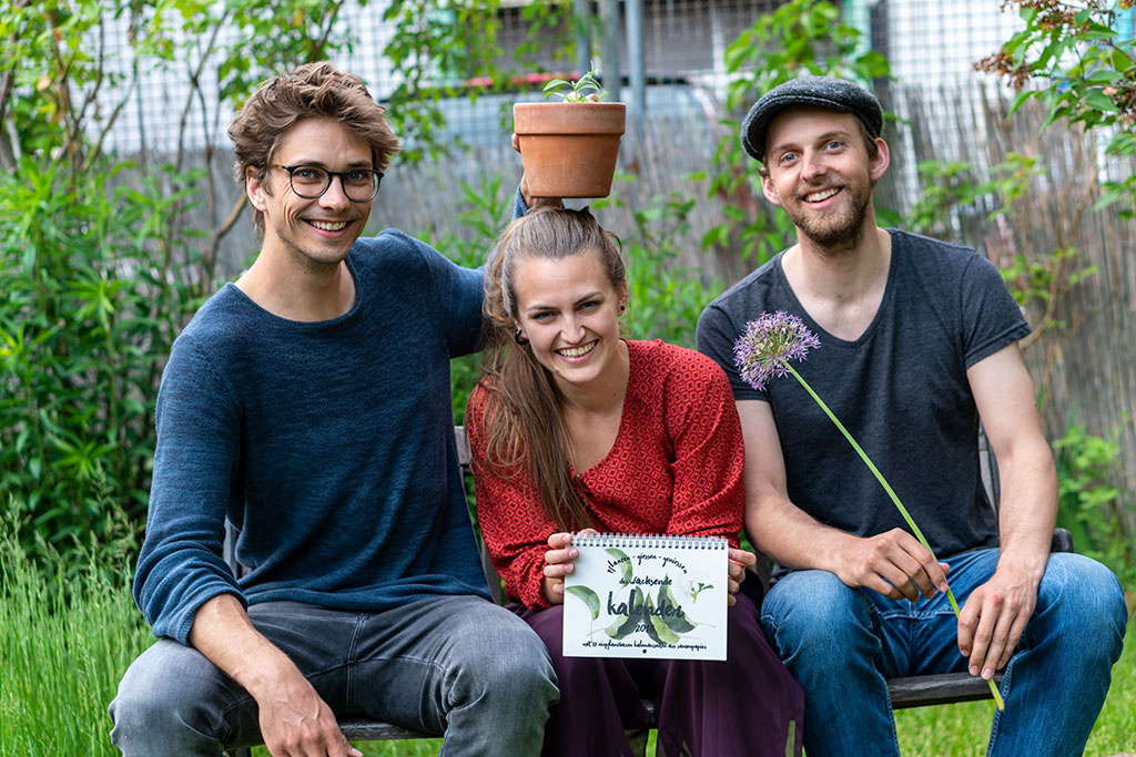 v.l.n.r.: Orlando Zaddach, Manuela Baron und Tobias Aufenanger sitzen in der Hocke und halten über Manuela einen Blumentopf.