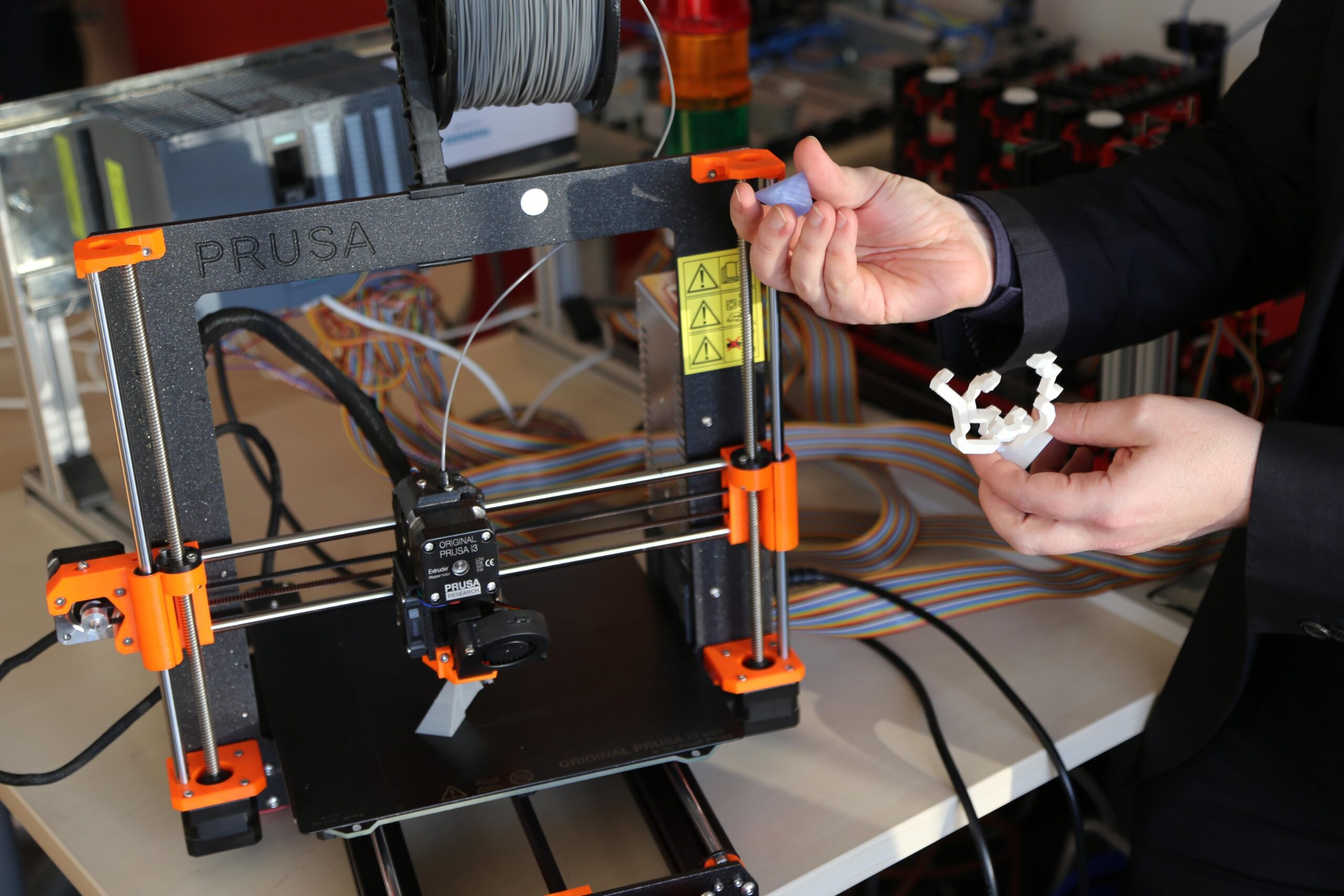 Es ist ein 3D-Drucker zu sehen. Rechts im Bild zeigt jemand kleine Gegenstände, die gedruckt wurden.