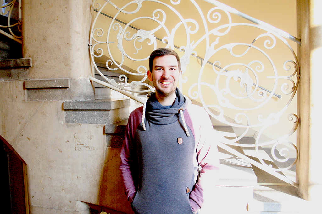 Portraitbild des Kurators: Er steht vor einem verschnörkeltem Treppengeländer, das von der rechten Seite mit Sonne angestrahlt wird.