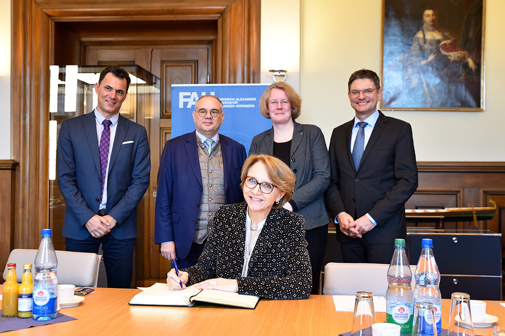 Die französische Botschaferin sitzt vor einem Buch und blickt in die Kamera. Im Hintergrund stehen vier weitere Personen, die ebenfalls in die Kamera blicken.