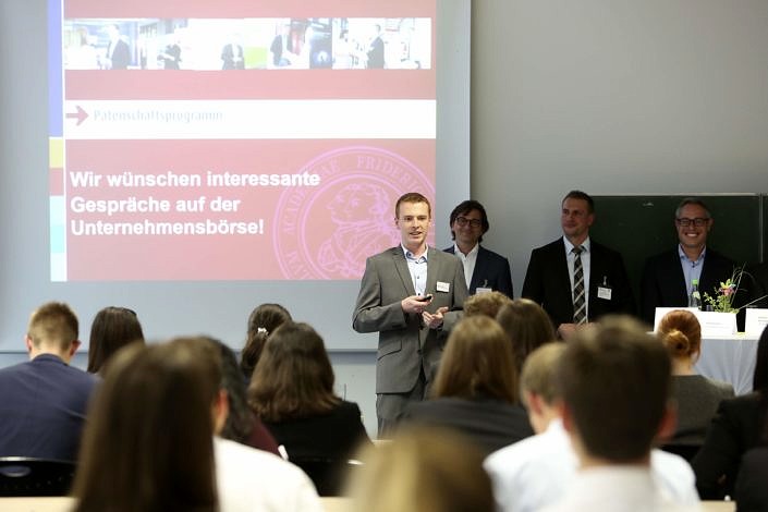 Ein Mann spricht in ein Mikrofon. Im Hintergrund ist eine Powerpoint-Präsentation zu sehen sowie weitere Männer in Anzügen.