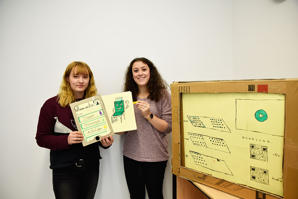 Zwei Studentinnen halten eine aus Karton gebastelte App-Simulation in die Höhe.