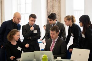 Studierende in Anzügen diskutieren vor Laptops.