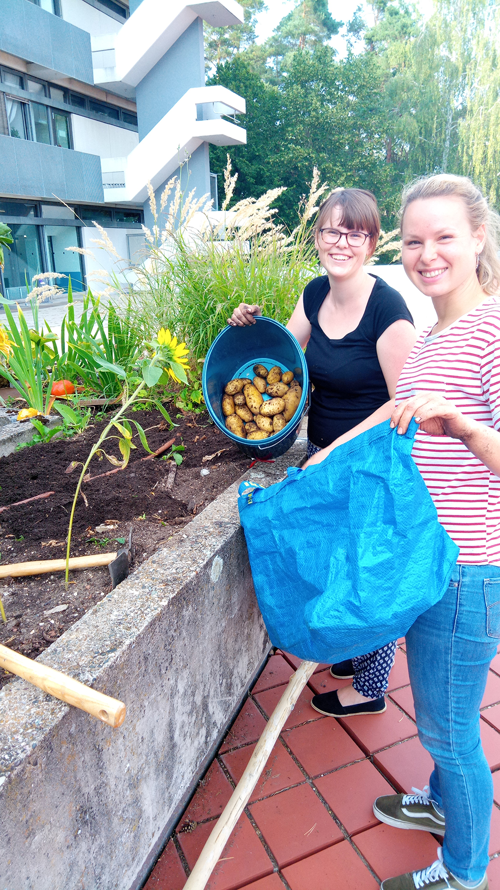 Eine Frau zeigt einen Eimer, in dem Kartoffeln sind und hält in der anderen Hand eine blaue Tüte.