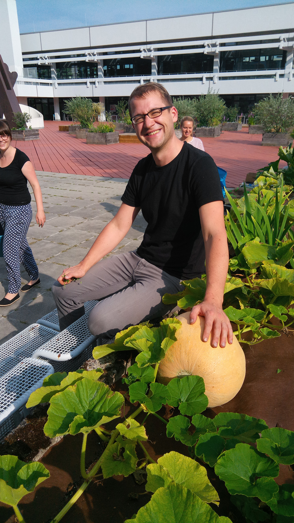 Ein Mann sitzt auf einer Mauer und hat seine Hand auf einen großen Kürbis gelegt, neben dem sich auch noch Salat und andere Gemüsesorten befinden.