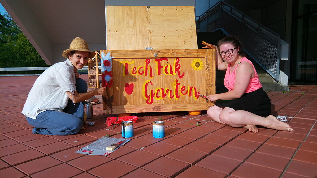 Zwei Frauen sitzen mit Pinsel und Farbe neben einer Holzkiste und blicken in die Kamera. Auf der Holzkiste steht in gelber und roter Farbe 