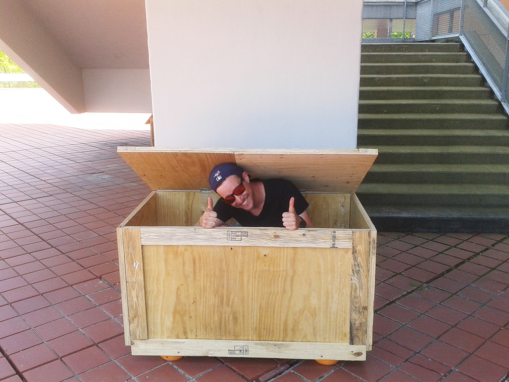 Ein Mann mit umgedrehter Cappy und Sonnenbrille sitzt in einer Holzkiste, schaut heraus und zeigt beide Daumen hoch.