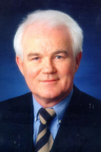 Portraitbild eines älteren Mannes mit weißem Haar im Anzug vor einem blauen Hintergrund