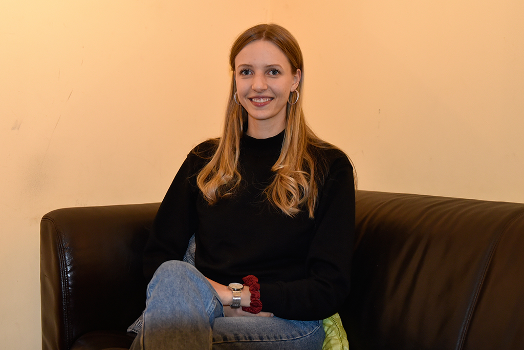 Eine Frau mit langen blonden Haaren sitzt auf einer schwarzen Ledercouch und blickt in die Kamera. Der Hintergrund wirkt warm orange.