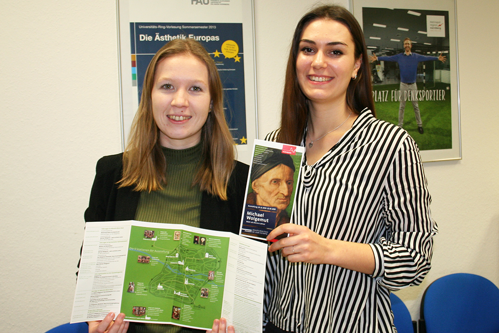 Zwei junge Frauen stehen nebeneinander, lächeln in die Kamera und zeigen Flyer.