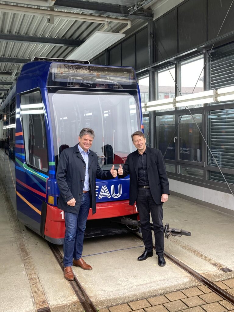 FAU-Präsident Prof. Dr. Joachim Hornegger und Gerd Lamatsch von der Stadtreklame Nürnberg GmbH zeigen Daumen hoch vor der Straßenbahn