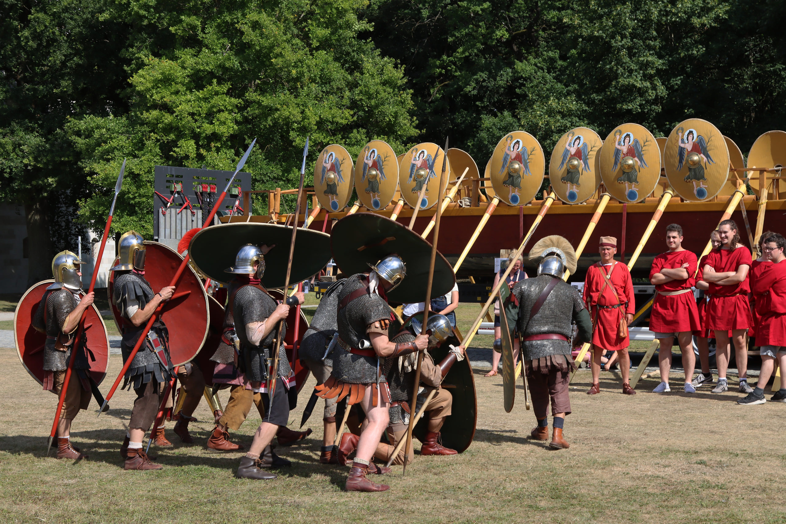 Reenactment-Darsteller in römischer Soldatenuniform stellen einen Schildwall auf. im Hintergrund steht ein Teil der Mannschaft eines Römerbootes in roten Gewändern.