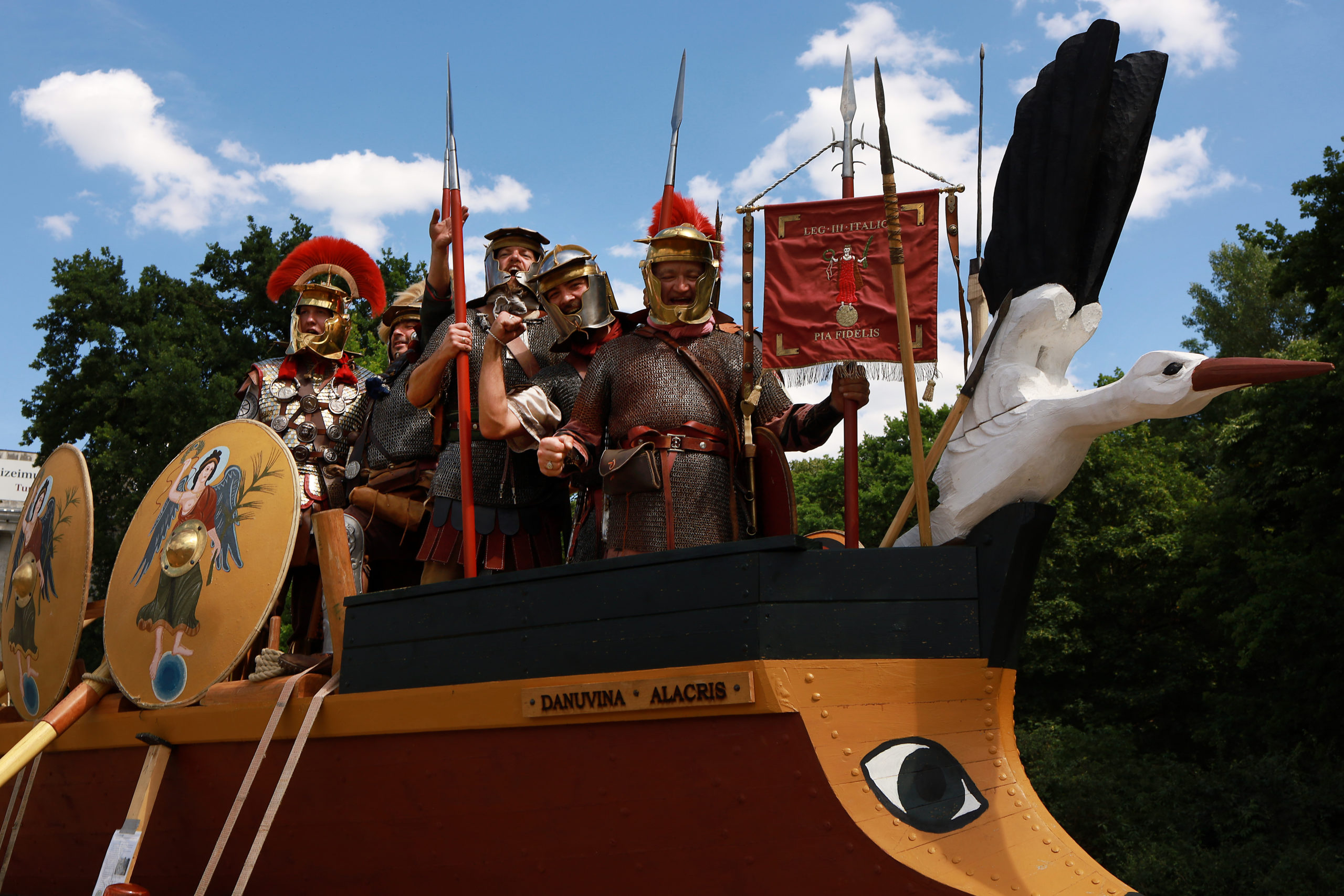 Fünf Männer in authentischen römischen Legionärsuniformen stehen im Bug eines Römerbootes und jubeln.