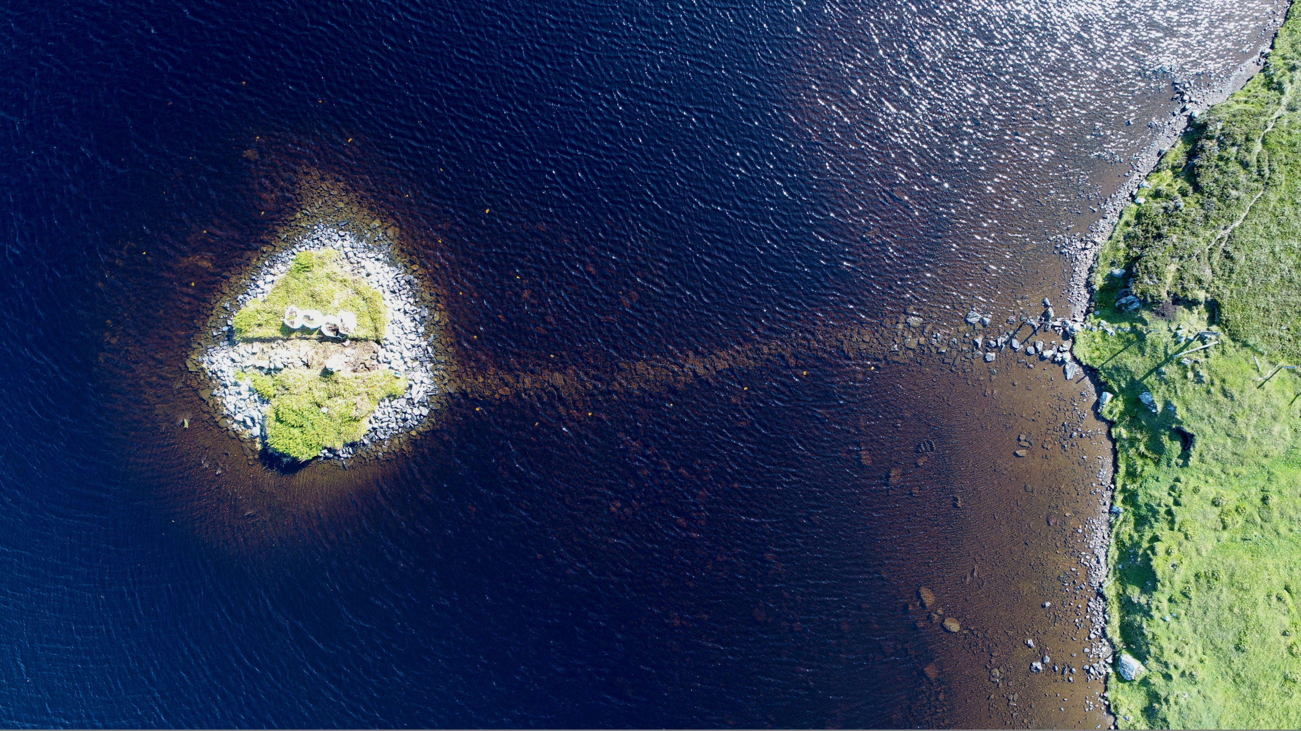 Luftaufnahme eines Sees mit Crannog. Durch das dunkelblaue Wasser ist eine Art Weg zu sehen, der zum Crannog führt.