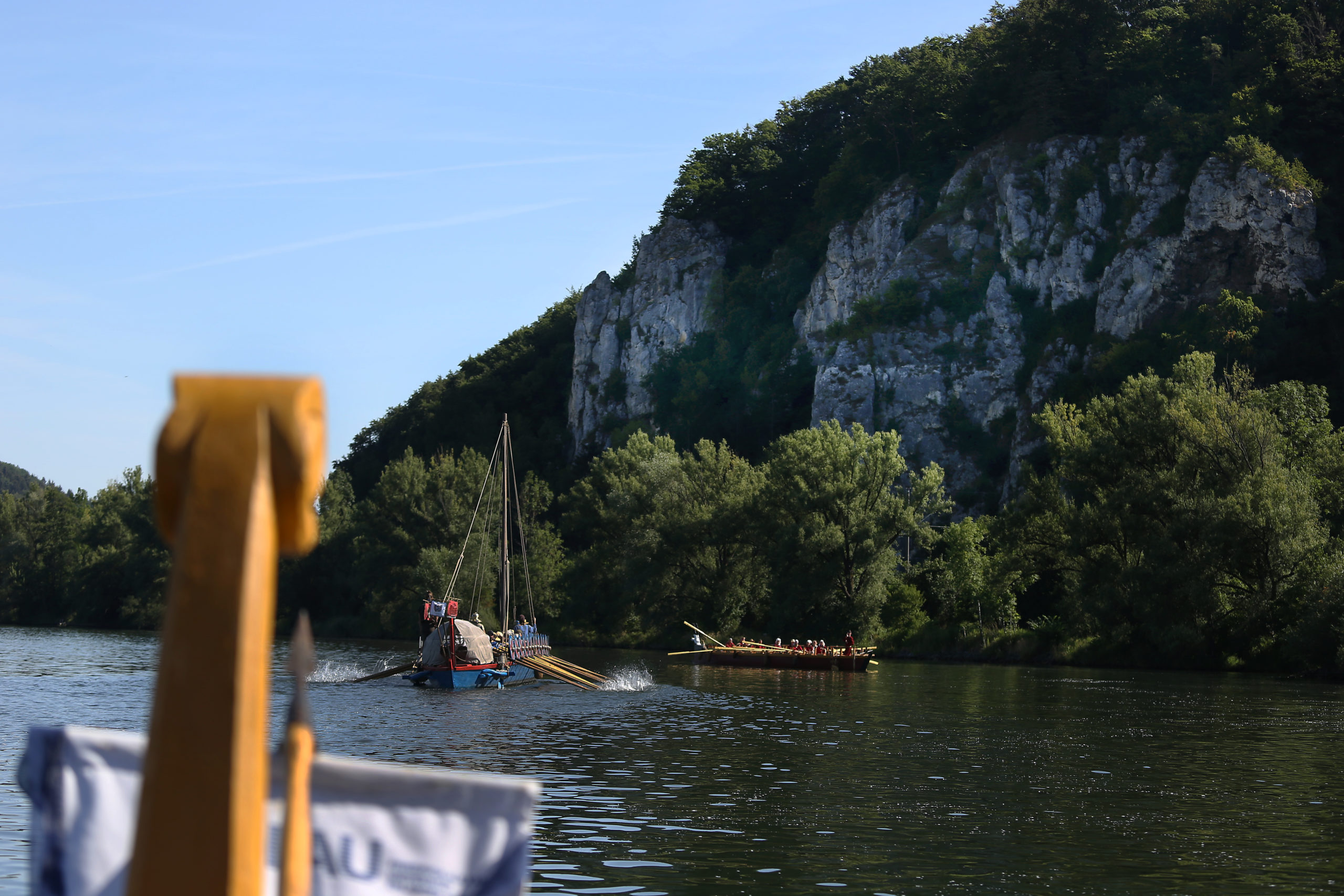 Vor einem großen Felsen schwimmen drei Römerboote in der Donau.