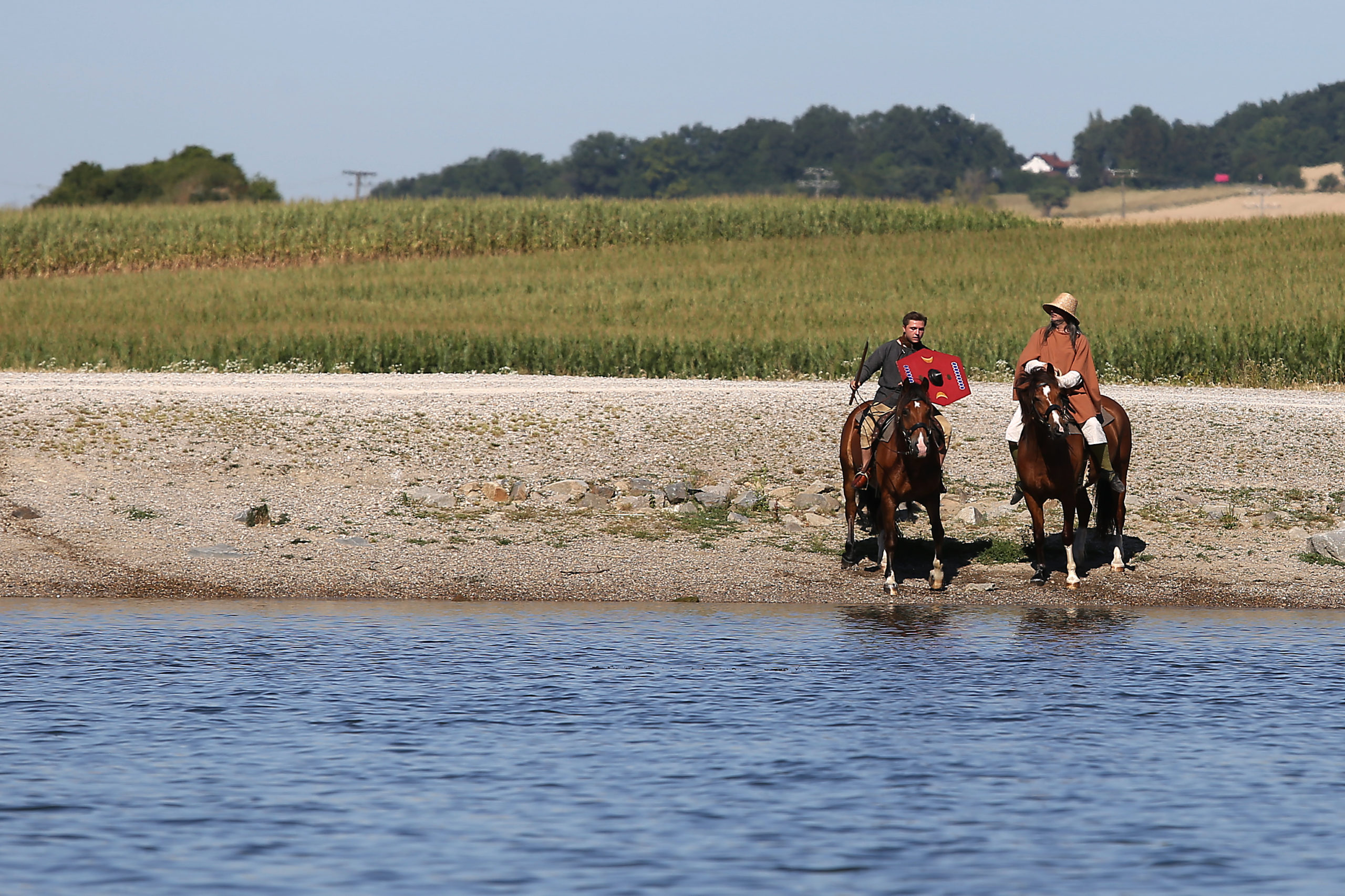 An einem Kiesstrand stehen zwei Pferde. Auf ihnen reiten zwei Reeanactment Darsteller.