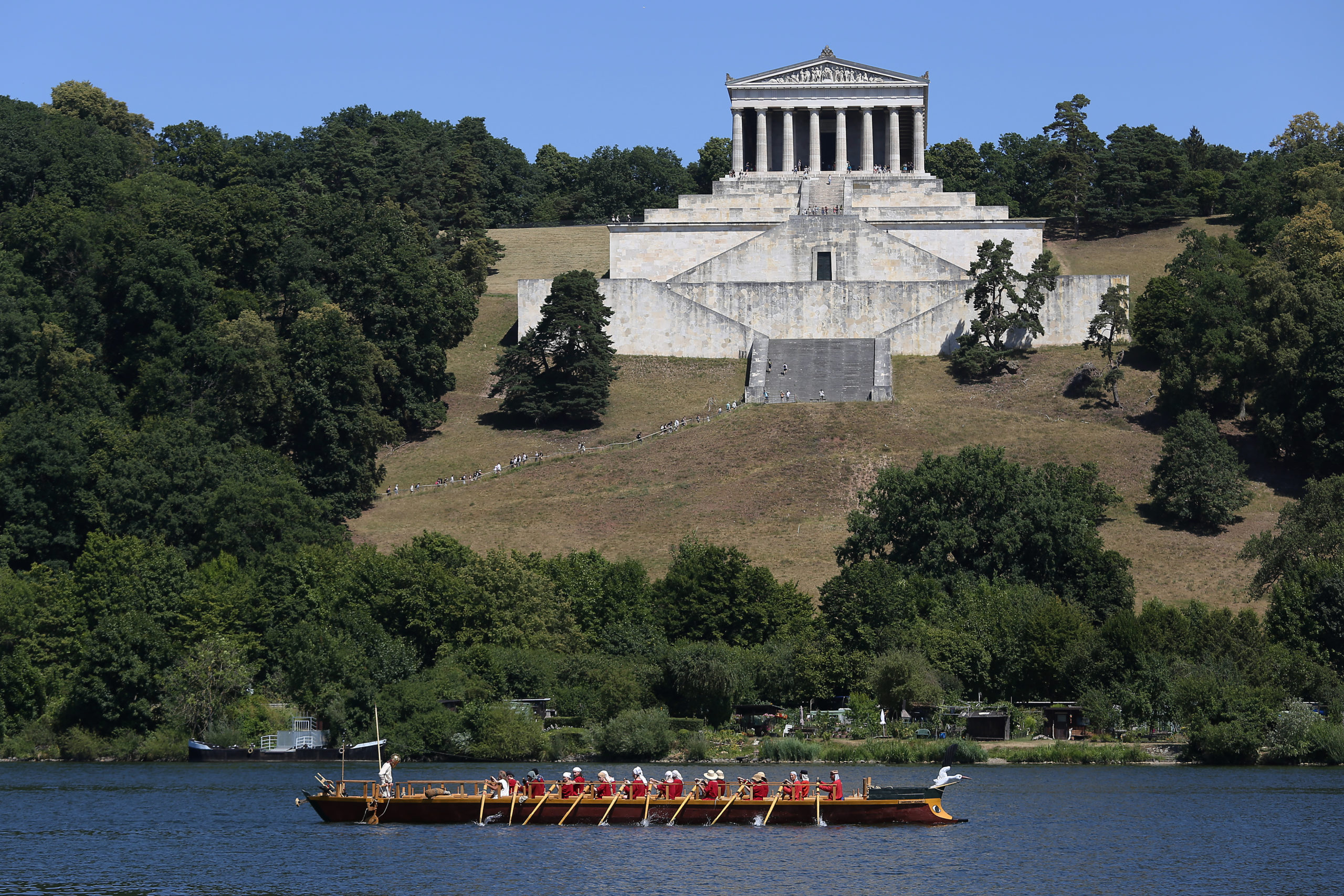 Im Vordergrund ist das Römerboot zu sehen, das auf der Donau schwimmt. Mehrere rotgekleidete Menschen rudern das Boot. Im Hintergrund sieht man die Walhalla.
