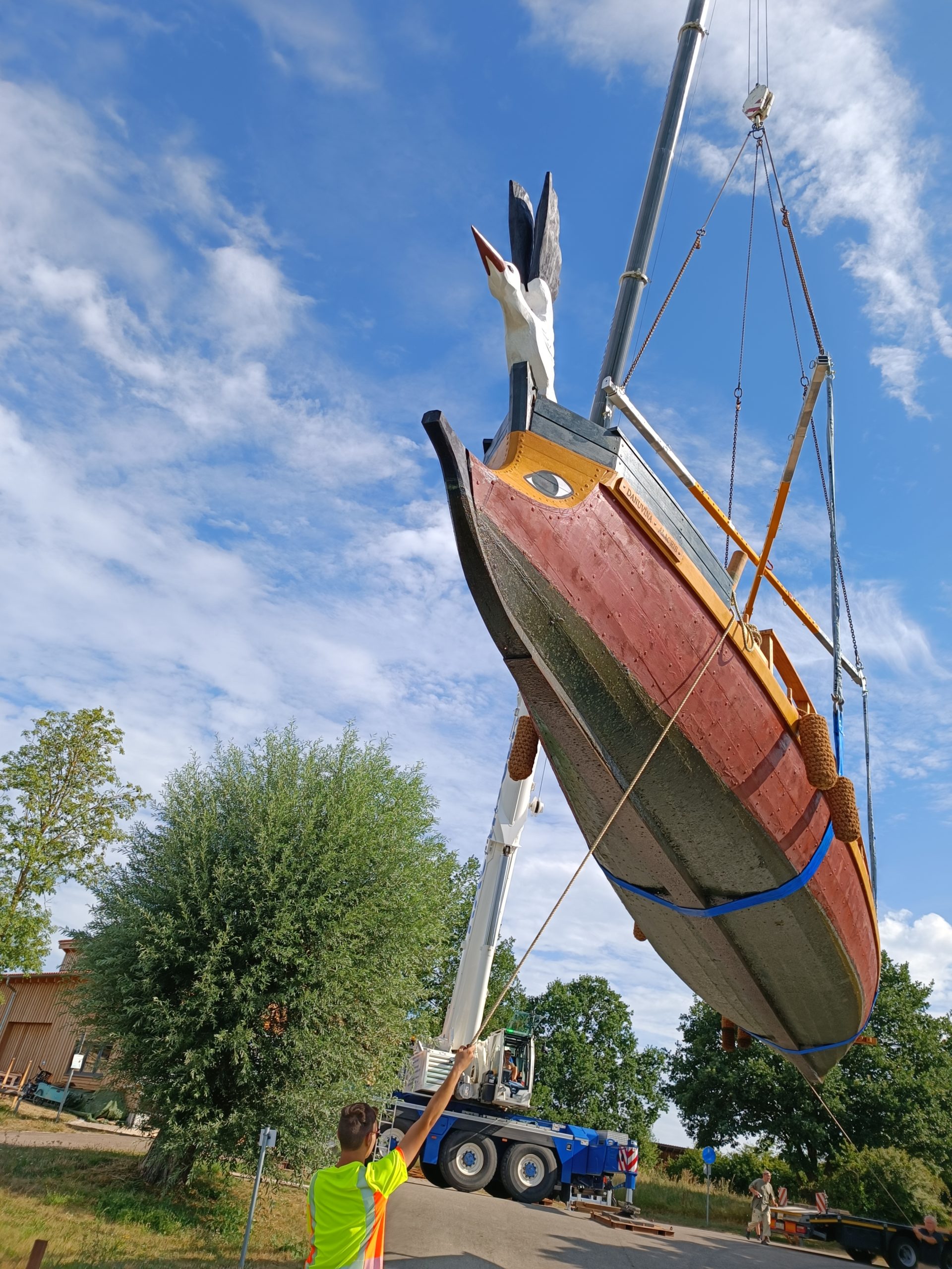 Ein Römerboot hängt an einem Kran. Ein Mann in greller Kleidung führt das Boot mit einem Seil.