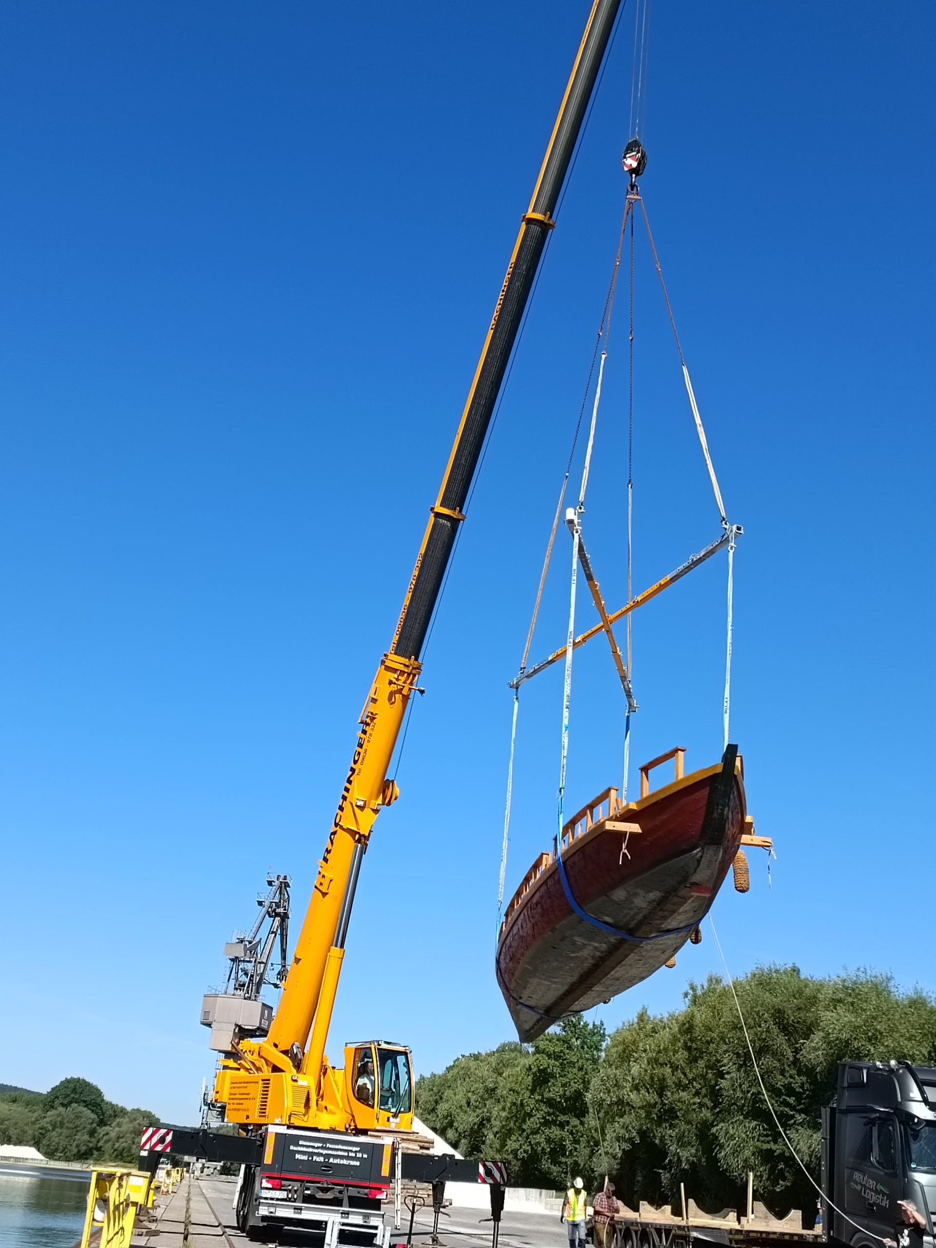 Ein Kran hebt ein Römerboot vom LKW hoch. Der Kran steht direkt neben dem Wasser.