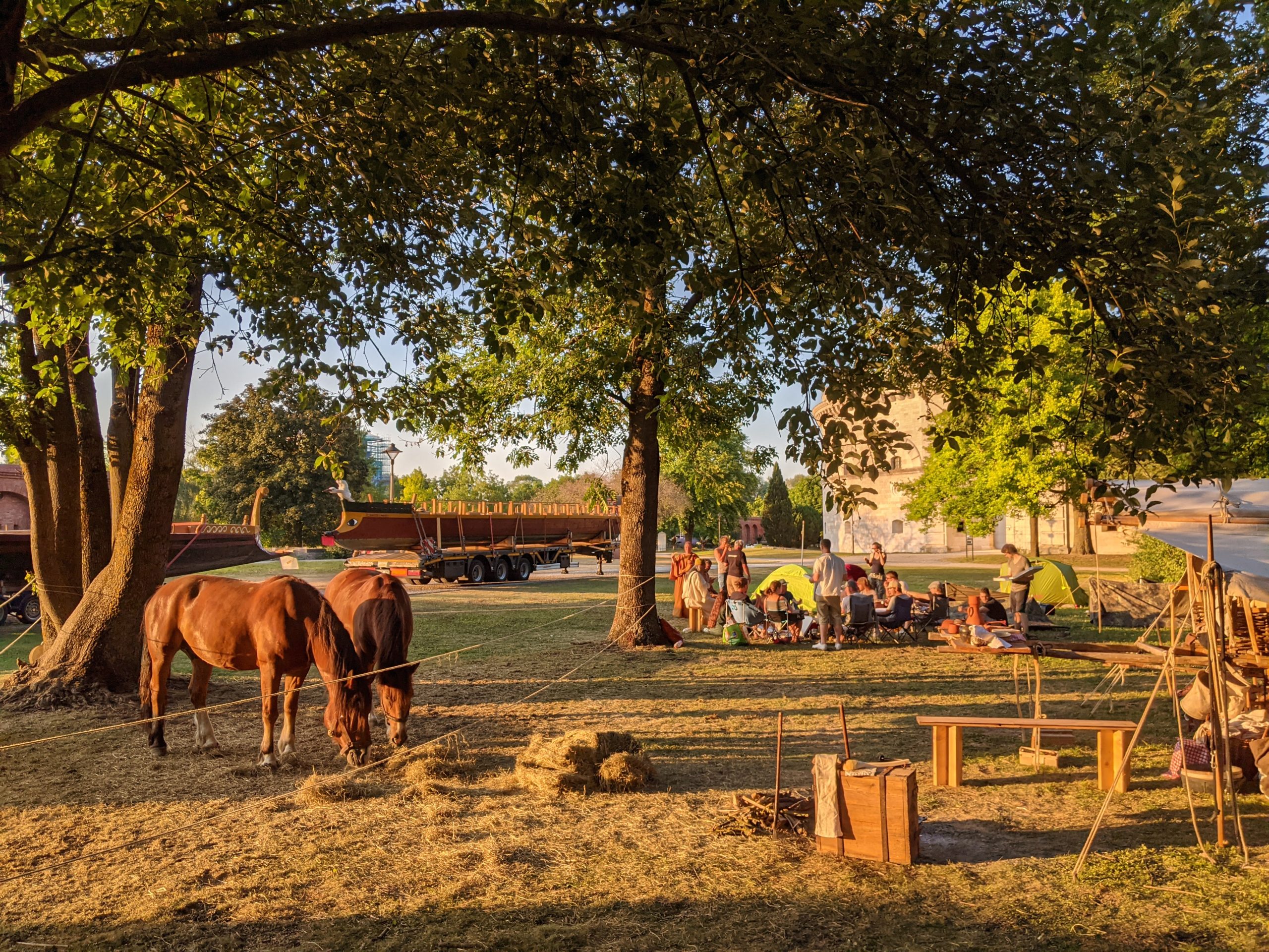 Im Abendrot macht eine Reenactmentgruppe Pause. Auf einer kleinen Koppel grasen zwei Pferde. Im Hintergrund stehen zwei Römerboote auf LKW-Anhängern.