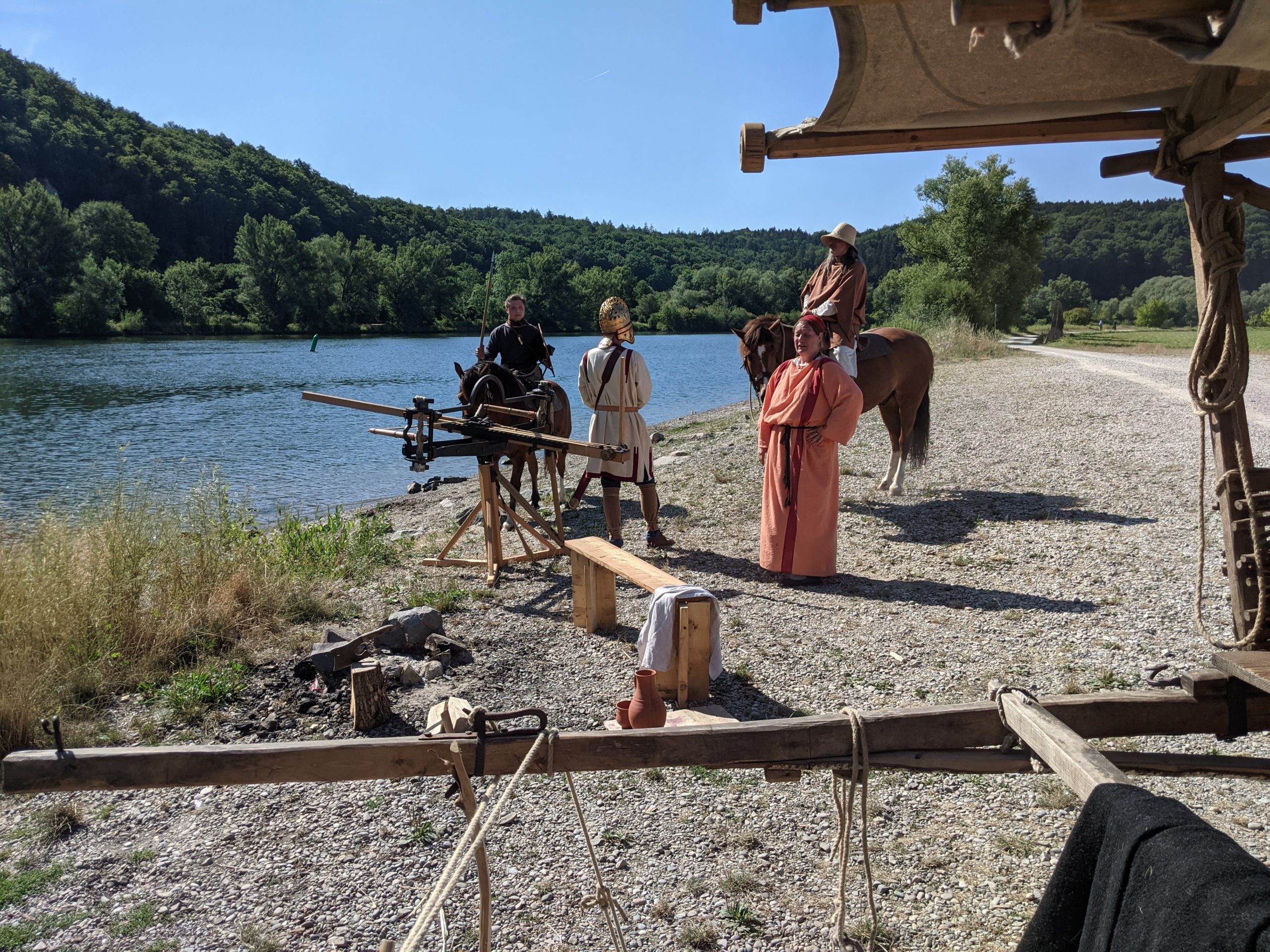 Am Ufer stehen vier Reenactment Darstellerinnen und Darsteller. Zwei der Personen reiten auf Pferden.