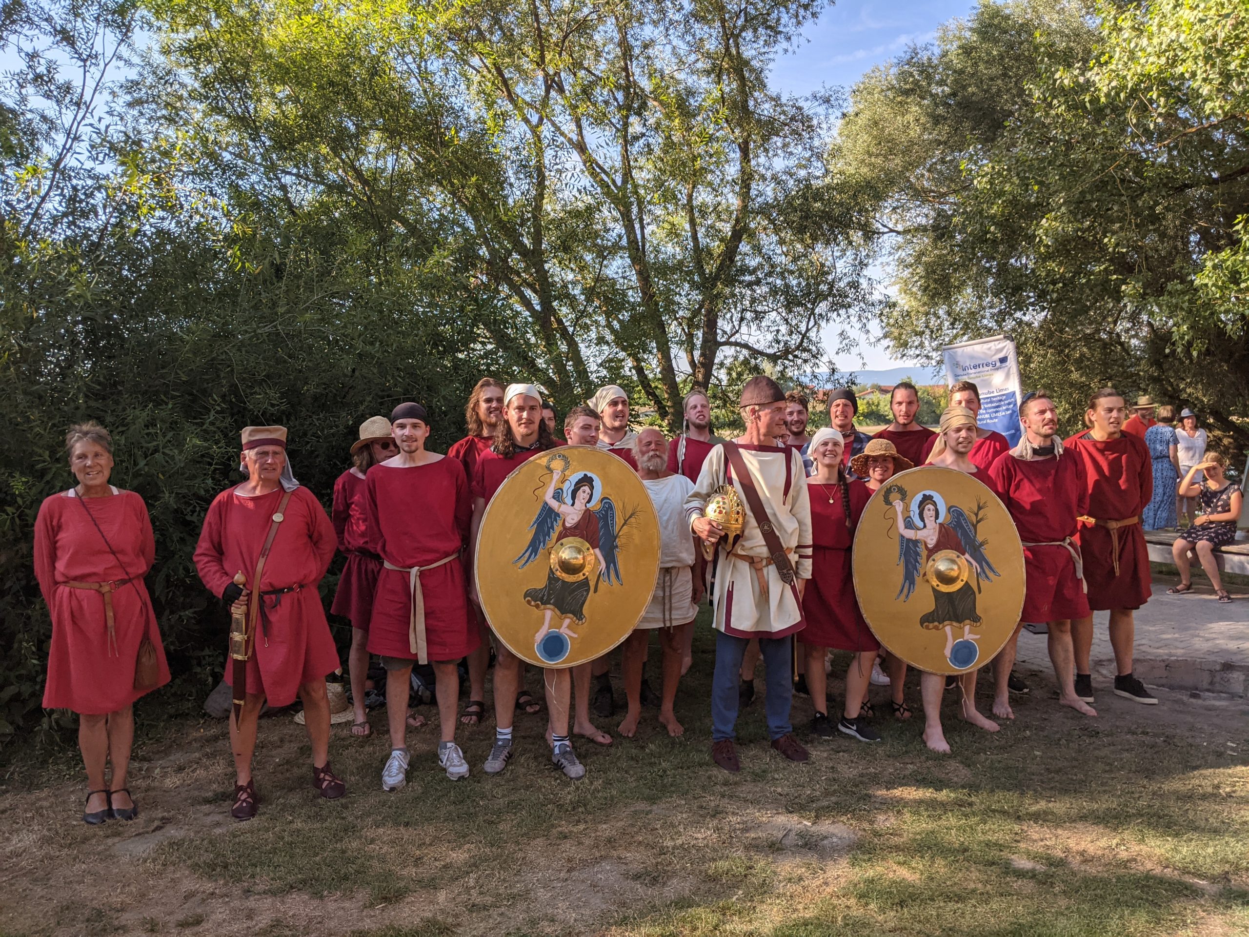 Gruppenfoto: Männer und Frauen in roten und weißen Gewändern halten große gelbe Schilde.