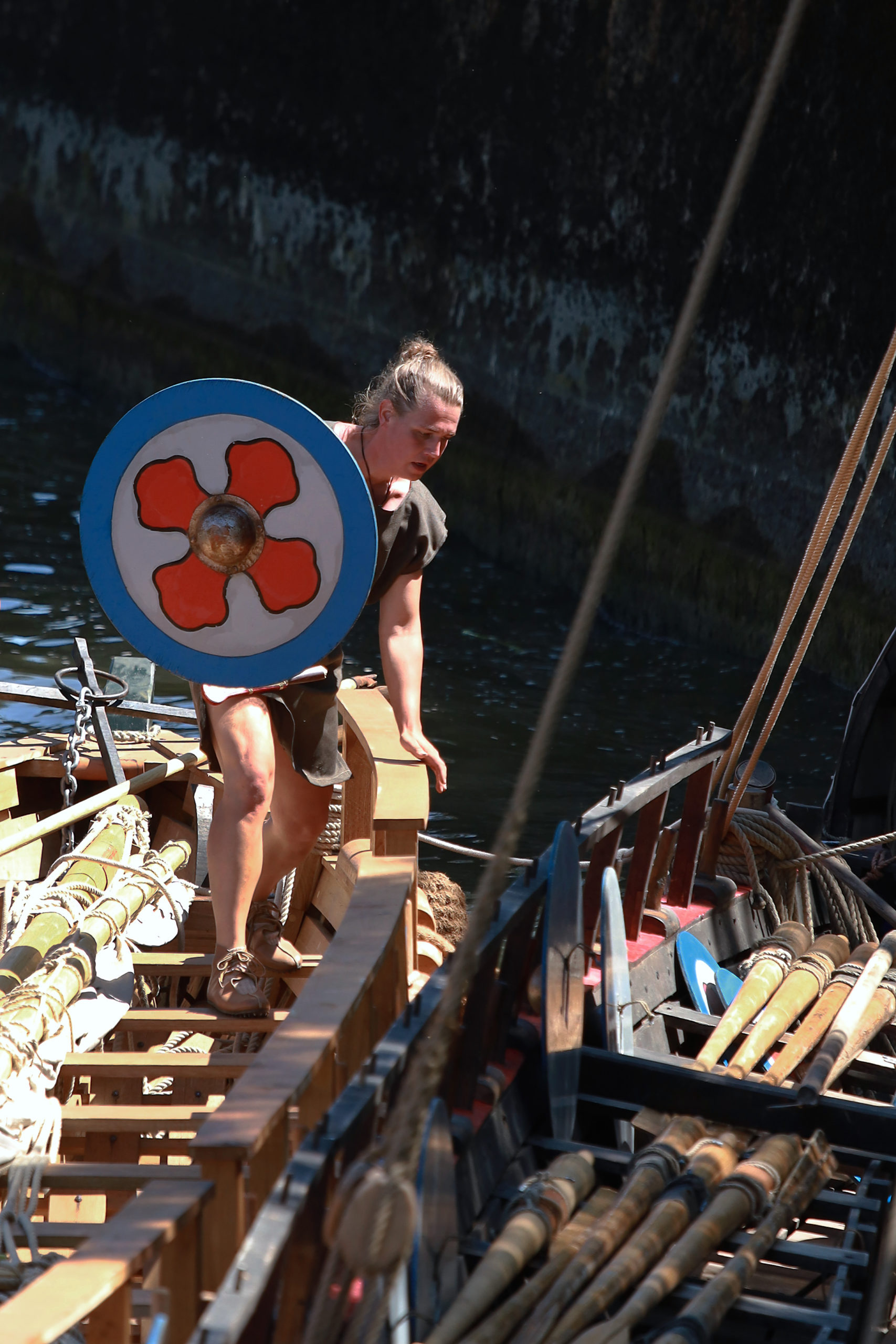 Ein Mann mit ein Schild klettert über die Sitzbänke eines Römerbootes.