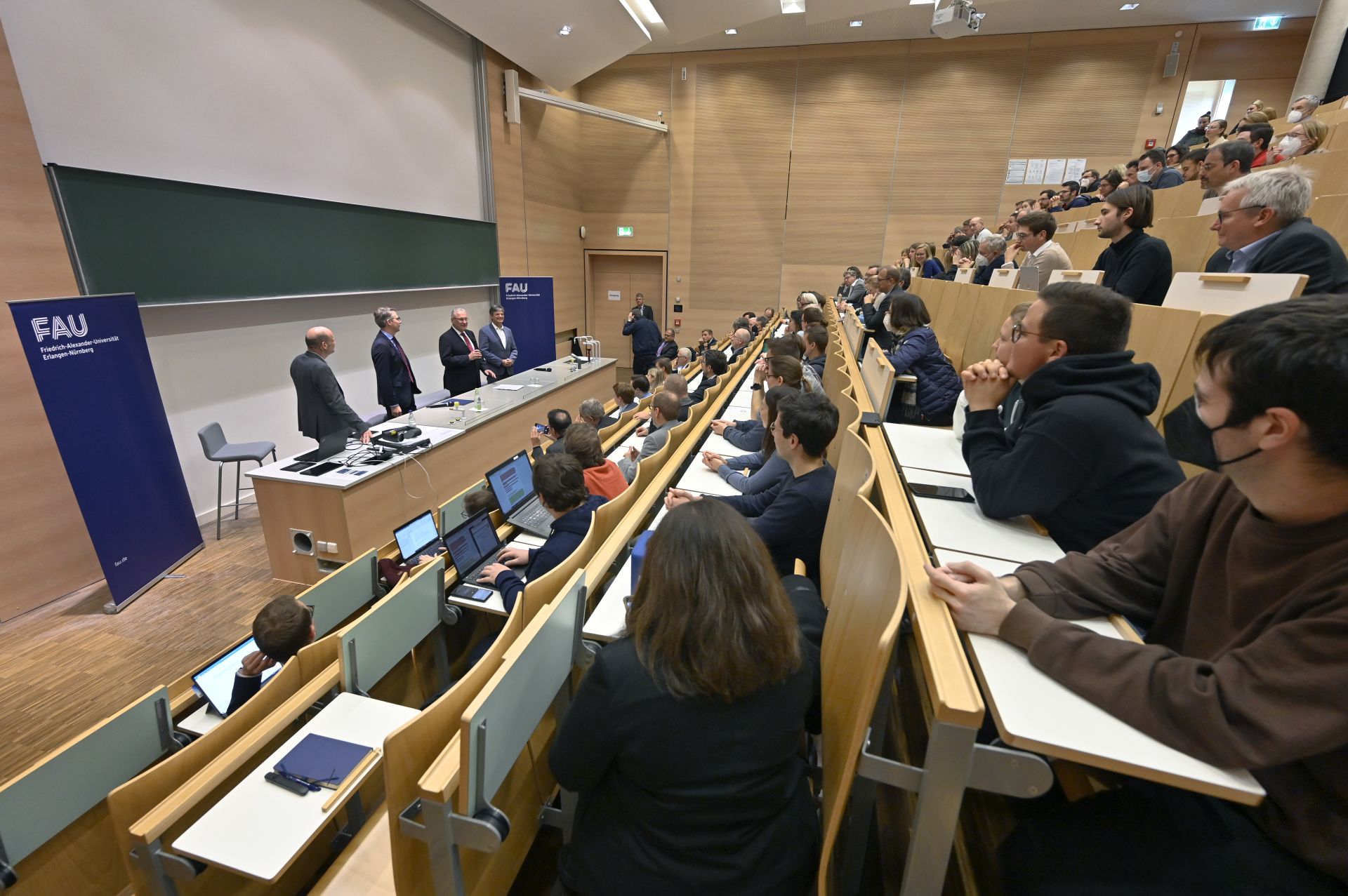 Blick in den Hörsaal über die Köpfe der Zuschauerinnen und Zuschauer auf die Bühne, wo vier Männer stehen.