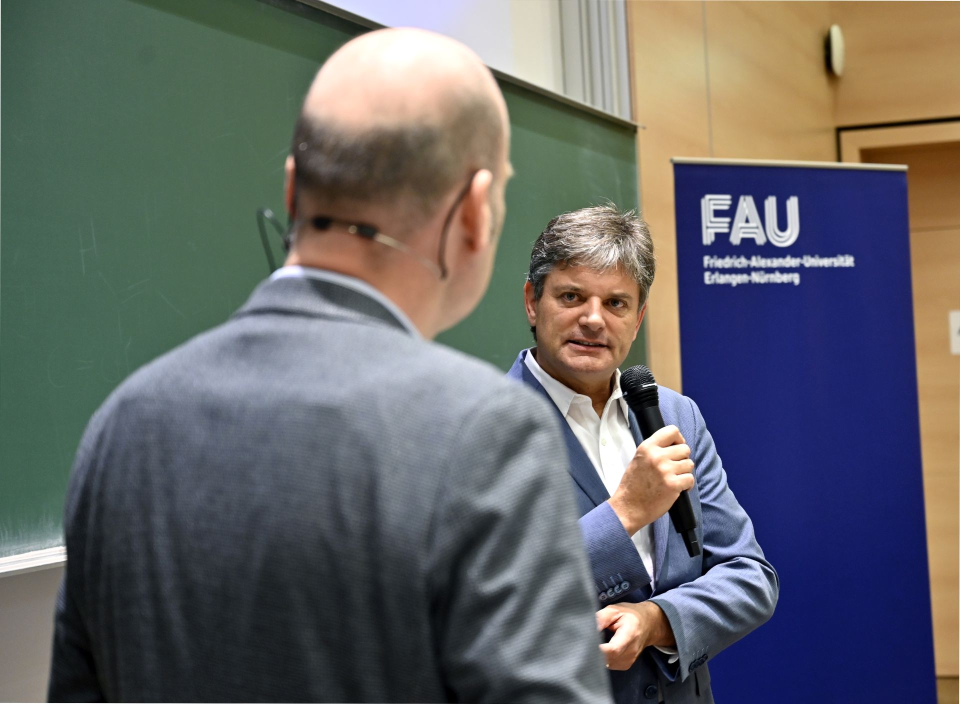Zwei Männer im Gespräch. Das Foto wurde über die Schulter des einen Mannes gemacht. Der zweite Mann hält ein Mirko in der Hand.