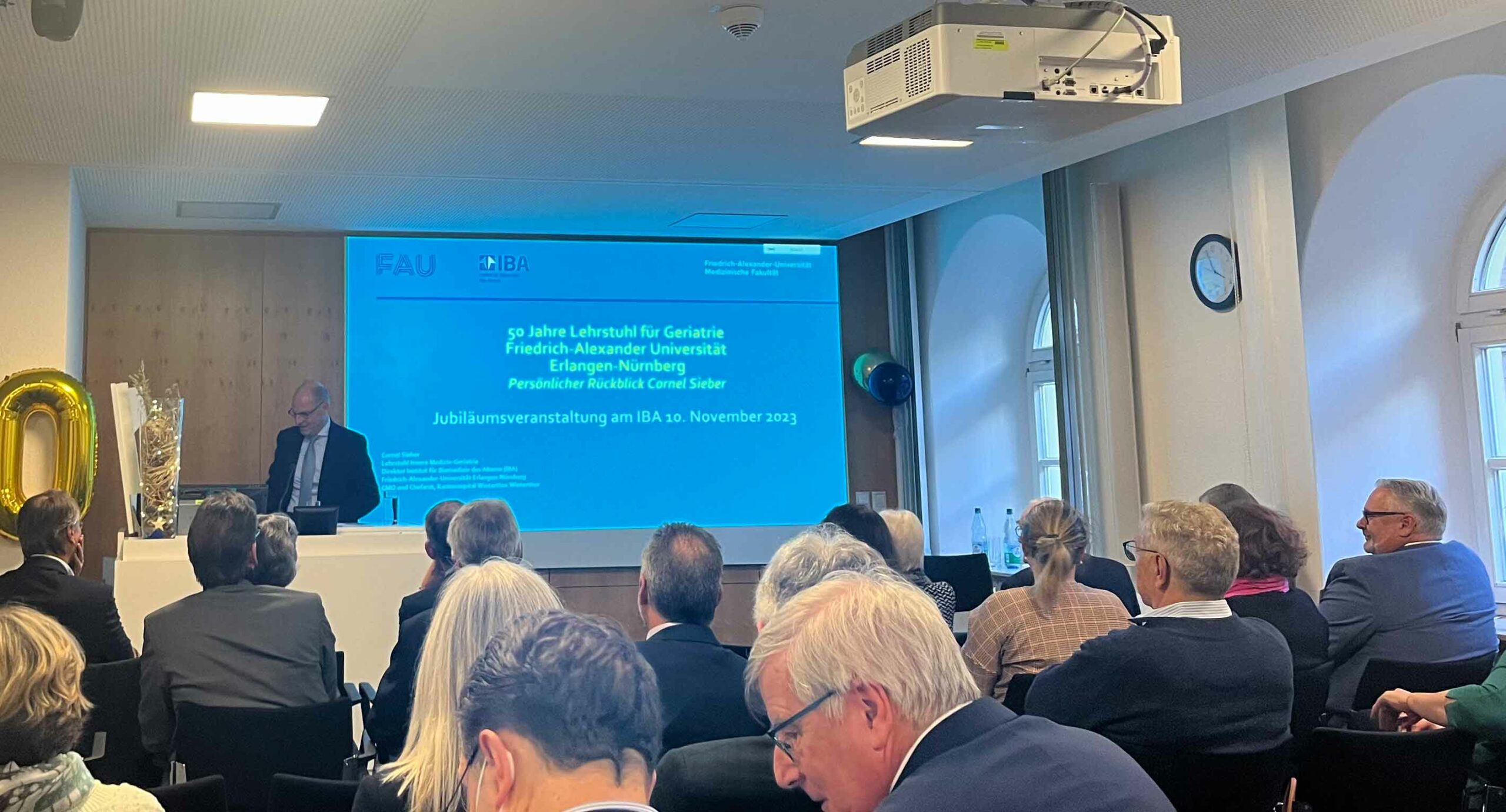Menschen sitzen vor Leinwand, auf der ein persönlicher Rückblick von Cornel Sieber angekündigt wird