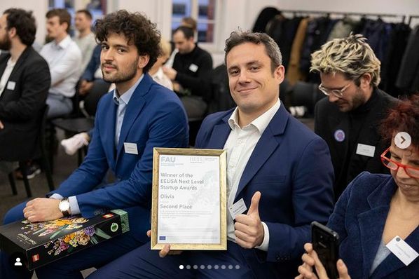 Zwei Männer in Bildmittelpunkt in Menge aus Menschen auf Stühlen, einer hält eine EELISA Urkunde und zeigt den Daumen hoch