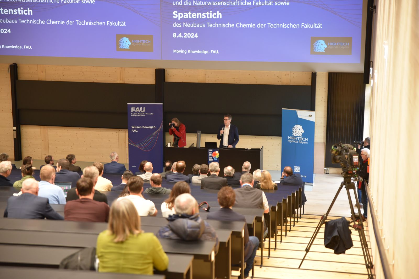 Hörsaal von hinten in den Raum aufgenommen. Man sieht die Treppe und Sitze absteigend nach unten sowie eine Projektion auf der Stirnseite des Saals.